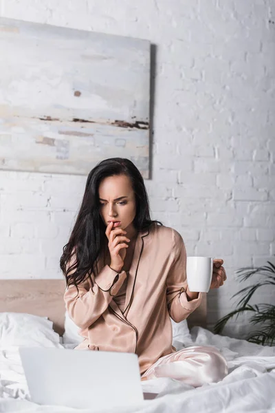 Worried young brunette woman sitting in bed with mug and laptop at morning — Stock Photo