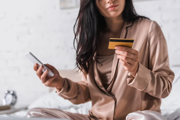 Vista recortada de la joven morena sentada en la cama con teléfono inteligente, tarjeta de crédito por la mañana - foto de stock