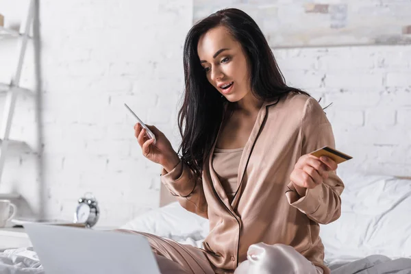 Choquée jeune femme brune assise au lit avec smartphone, carte de crédit et ordinateur portable le matin — Photo de stock