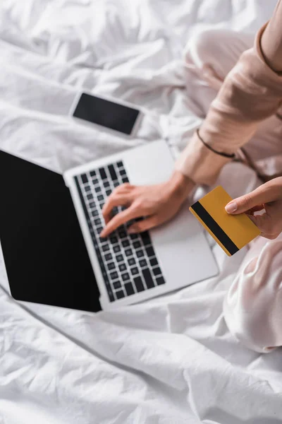 Vista recortada de la mujer sentada en la cama con teléfono inteligente, tarjeta de crédito y portátil por la mañana - foto de stock