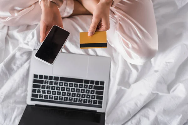 Vista cortada da mulher sentada na cama com smartphone, cartão de crédito e laptop pela manhã — Fotografia de Stock