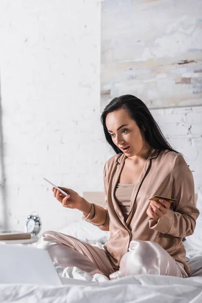 Giovane donna bruna scioccata seduta a letto con smartphone, carta di credito e laptop al mattino — Foto stock