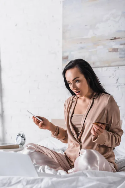 Choquée jeune femme brune assise au lit avec smartphone, carte de crédit et ordinateur portable le matin — Photo de stock