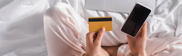 Vue recadrée de la femme assise au lit avec smartphone, carte de crédit et ordinateur portable le matin, bannière — Photo de stock