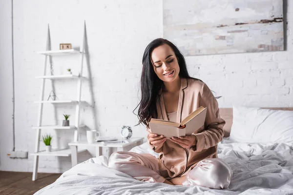 Sorridente giovane donna bruna seduta a letto con libro al mattino — Foto stock