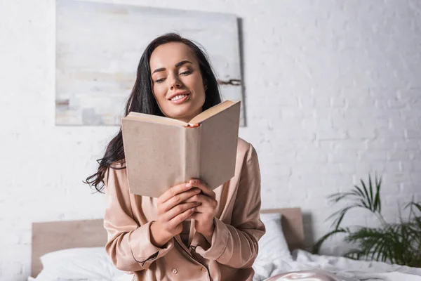 Sonriente joven morena sentada en la cama con libro por la mañana - foto de stock