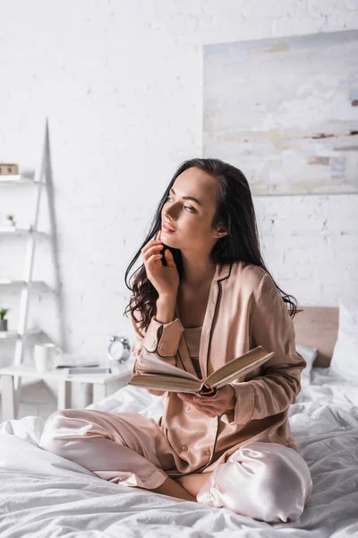 Mujer morena joven soñadora sentada en la cama con libro por la mañana - foto de stock