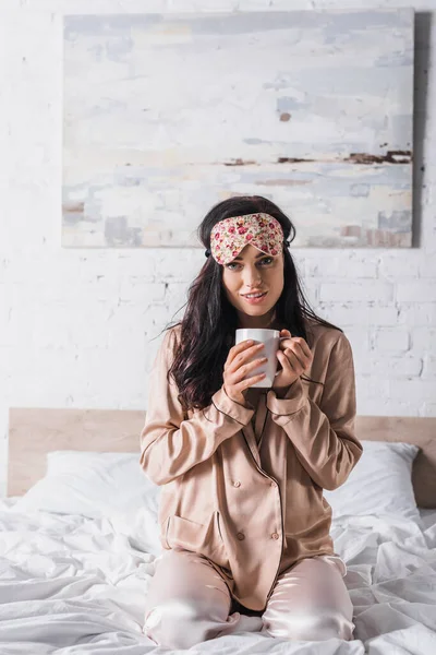 Young brunette woman sitting in bed in sleep mask with mug of cocoa — Stock Photo