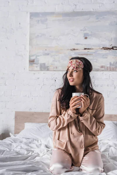 Young brunette woman sitting in bed in sleep mask with mug of cocoa — Stock Photo