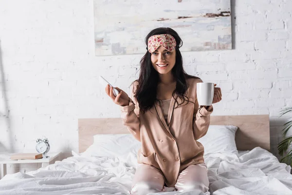 Улыбающаяся юная брюнетка сидит в постели в маске для сна с кружкой какао и смартфоном — стоковое фото