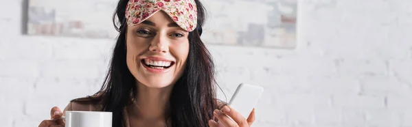 Giovane donna bruna sorridente in maschera del sonno con tazza di cacao e smartphone, banner — Foto stock