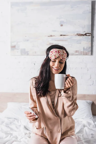 Sonriente joven morena sentada en la cama en la máscara del sueño con taza de cacao y teléfono inteligente - foto de stock