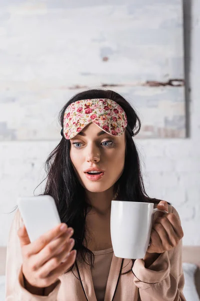 Shocked young brunette woman sitting in bed in sleep mask with mug of cocoa and smartphone — Stock Photo