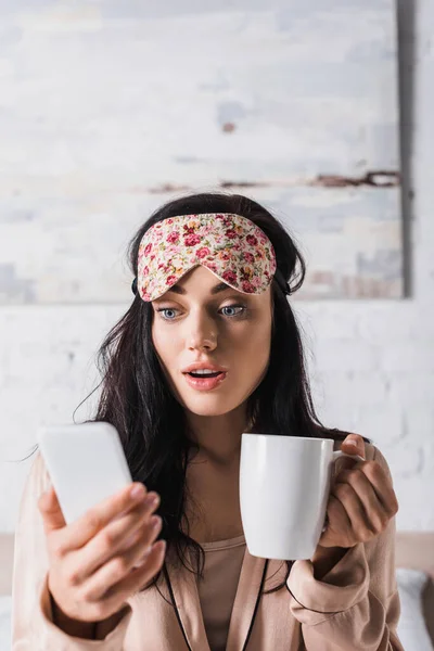 Scossa giovane donna bruna seduta a letto in maschera del sonno con tazza di cacao e smartphone — Foto stock