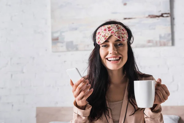 Felice giovane donna bruna seduta a letto in maschera del sonno con tazza di cacao e smartphone — Foto stock