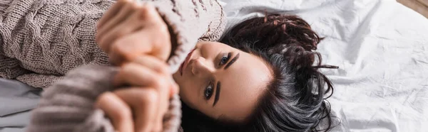 Vista superior de la joven morena en suéter y las manos extendidas borrosas acostado en la cama por la mañana, pancarta - foto de stock