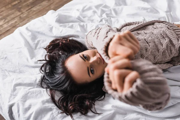 Top view of young brunette woman in sweater and blurred outstretched hands lying in bed at morning — Stock Photo