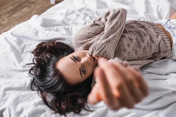 Top view of young brunette woman in sweater and blurred outstretched hand lying in bed at morning — стоковое фото