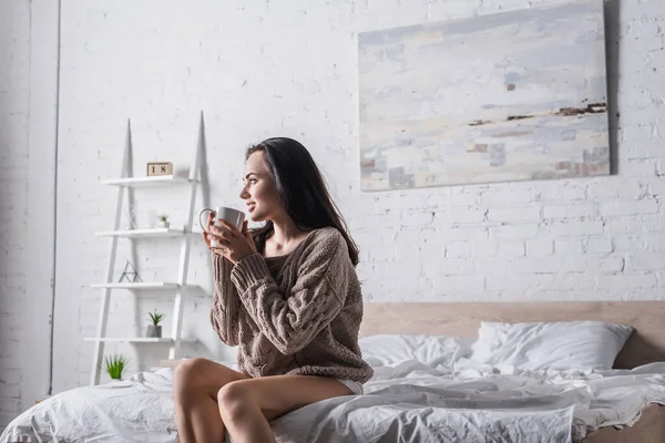 Junge brünette Frau im Pullover sitzt morgens mit einem Becher heißem Kakao im Bett — Stockfoto
