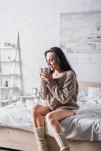 Jovem morena em suéter sentado na cama com caneca de cacau quente de manhã — Fotografia de Stock