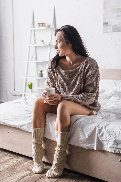 Jeune femme brune en pull et chaussettes assis au lit avec tasse de cacao chaud le matin — Photo de stock