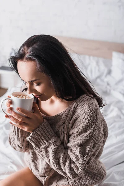 Giovane donna bruna in maglione seduta a letto con tazza di cioccolata calda al mattino — Foto stock