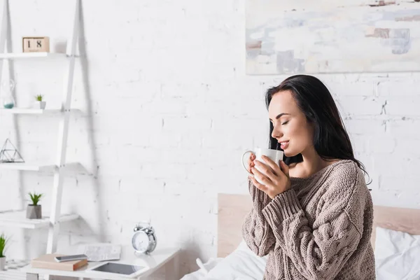 Joven morena mujer en suéter sentado en la cama con taza de cacao caliente por la mañana - foto de stock