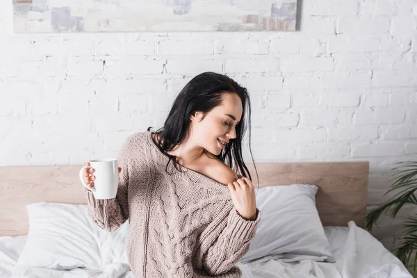 Jeune femme brune en pull avec épaule nue assise au lit avec tasse de cacao chaud le matin — Photo de stock