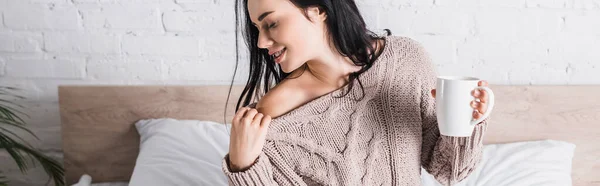 Giovane donna bruna in maglione con spalla nuda seduta a letto con tazza di cioccolata calda al mattino, striscione — Foto stock