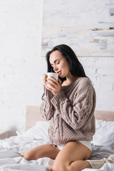 Сексуальная молодая брюнетка в свитере сидит в постели с кружкой горячего какао по утрам — стоковое фото