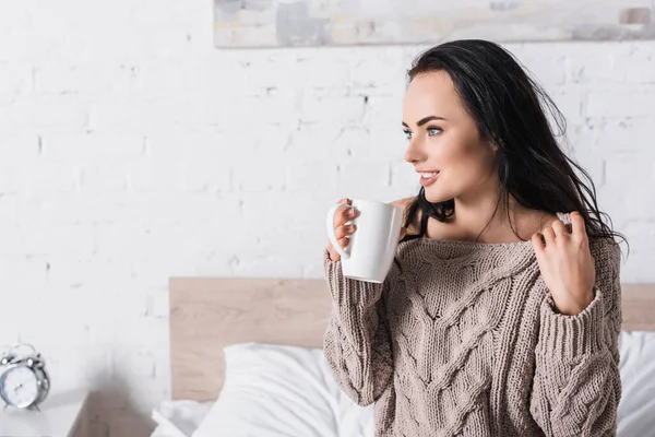 Jeune femme brune en pull assis dans le lit avec tasse de cacao chaud le matin — Photo de stock