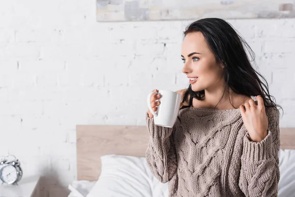 Junge brünette Frau im Pullover sitzt morgens mit einem Becher heißem Kakao im Bett — Stockfoto