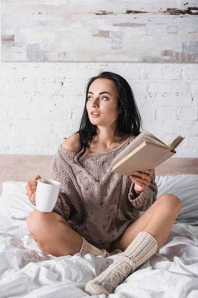 Mujer morena joven soñadora en suéter sentado en la cama con taza de cacao caliente y libro por la mañana - foto de stock