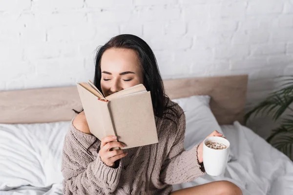 Glückliche junge brünette Frau im Pullover sitzt im Bett mit einem Becher heißen Kakaos und Buch am Morgen — Stockfoto