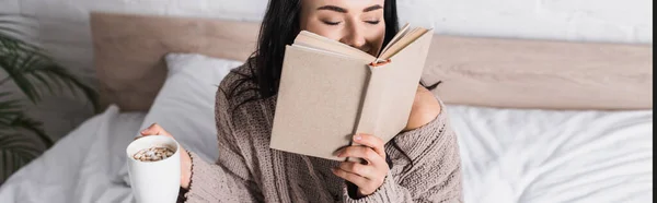Glückliche junge brünette Frau im Pullover sitzt im Bett mit einem Becher heißem Kakao und Buch am Morgen, Banner — Stockfoto