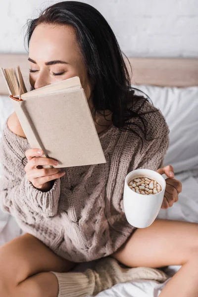 Молодая брюнетка в свитере сидит в постели с кружкой горячего какао и книга по утрам — стоковое фото