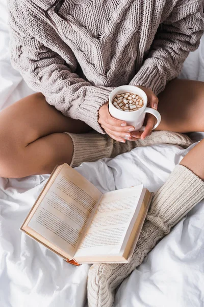Vue recadrée de la femme en pull et chaussettes tricotées assis dans le lit avec tasse de cacao chaud et livre le matin — Photo de stock