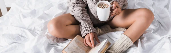 Abgeschnittene Ansicht einer Frau in Pullover und Stricksocken, die morgens mit einem Becher heißem Kakao und Buch im Bett sitzt, Banner — Stockfoto