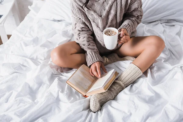 Vista cortada da mulher em suéter e meias de malha sentado na cama com caneca de cacau quente e livro de manhã — Stock Photo