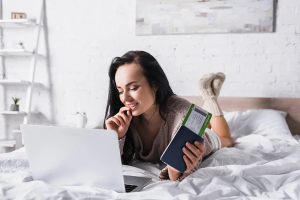 Lächelnde junge brünette Frau im Pullover mit Laptop und Flugticket im Bett liegend — Stockfoto