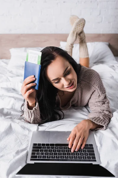 Jeune femme brune en pull couché dans le lit avec ordinateur portable et billet d'avion — Photo de stock