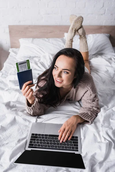 Счастливая юная брюнетка в свитере лежит в постели с ноутбуком и авиабилетом — стоковое фото