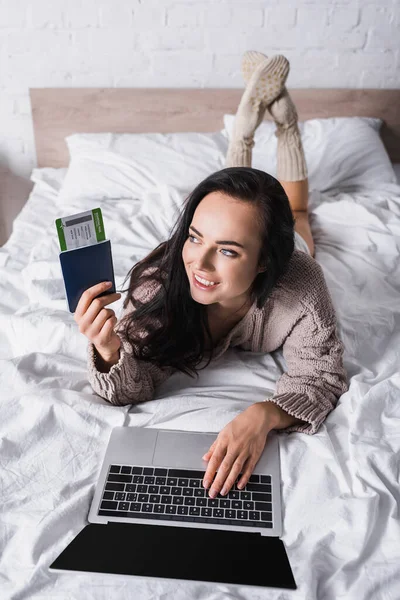 Happy young brunette woman in sweater lying in bed with laptop and air ticket — Stock Photo
