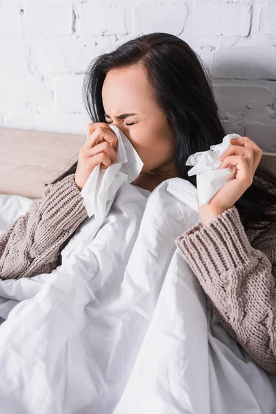Kranke junge brünette Frau im Pullover niest im Bett mit Gewebe — Stockfoto