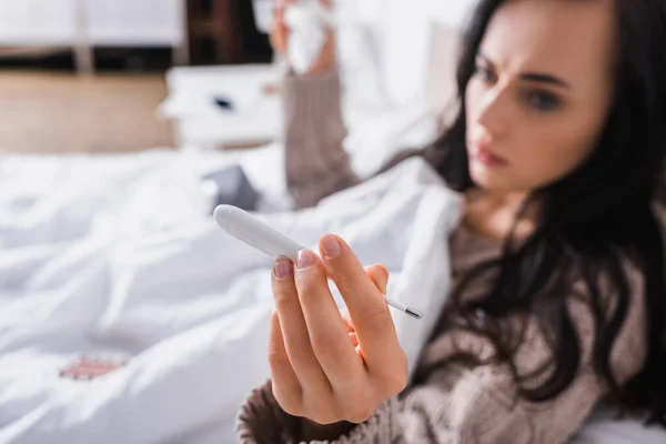 Offuscata malata giovane donna bruna in maglione sdraiata a letto con termometro e tessuto — Foto stock