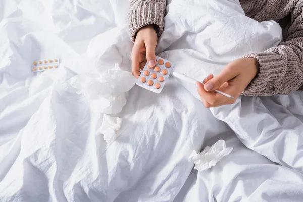 Vista ritagliata di giovane donna malata con tessuto, pillole e termometro a letto — Foto stock