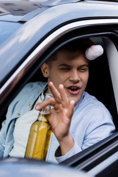 Borracho, hombre excitado en sombrero de santa sentado en el coche con botella de whisky en primer plano borrosa - foto de stock