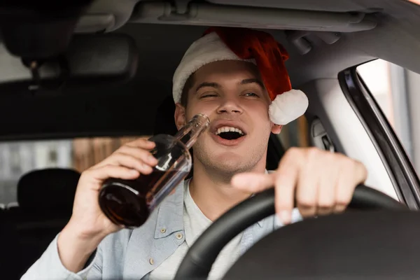 Betrunkener, aufgeregter Mann mit Weihnachtsmütze, der Auto fährt und eine Flasche Whiskey im verschwommenen Vordergrund hält — Stockfoto