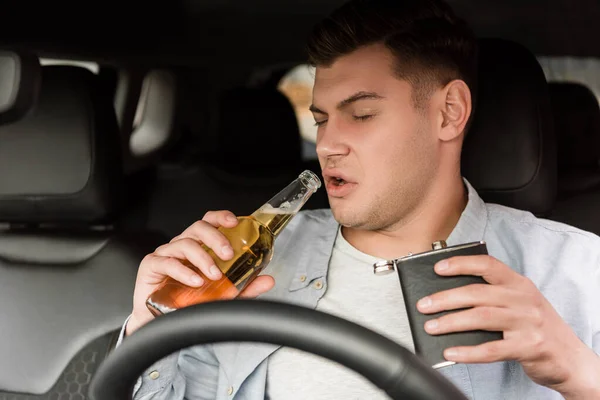 Drunk man holding flask and bottle of whiskey while sitting in car, blurred foreground — Stock Photo