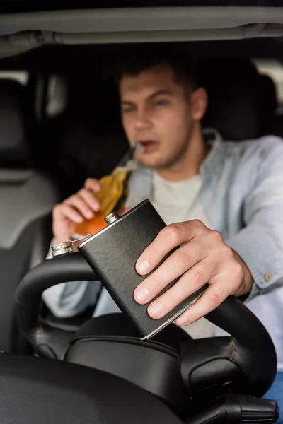 Young man holding flask and drinking whiskey while driving car, blurred background — Stock Photo